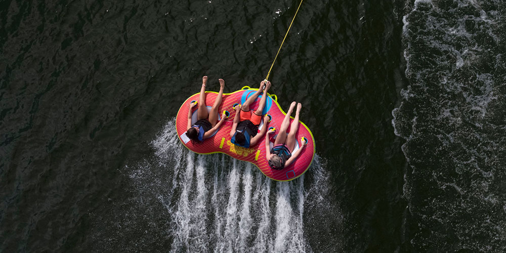 Aerial view of three people being pulled on a towable tube behind a boat on a lake
