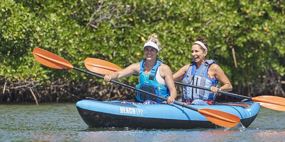 Two women approach a lake, pulling their kayaks behind them.