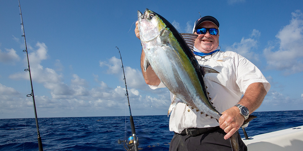Large, smiling man at stern of boat holding up big tuna
